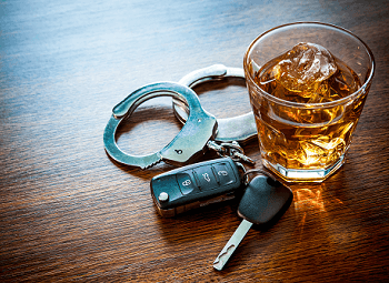 Glass of an alcoholic beverage with a pair of handcuffs and a set of car keys beside it on a wood table